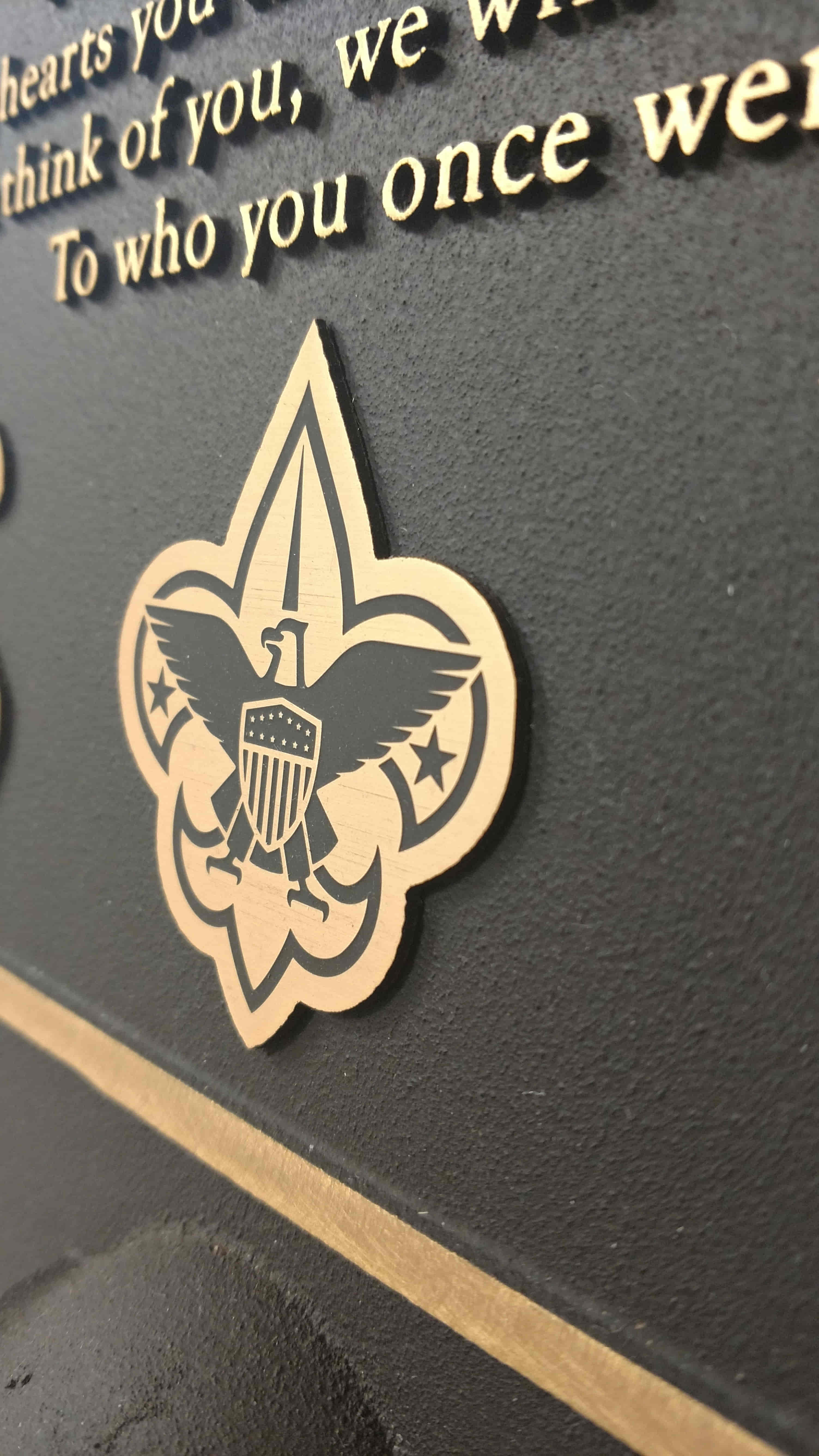 A close-up of a bronze emblem featuring an eagle and fleur-de-lis, with etched lettering and a textured black background.