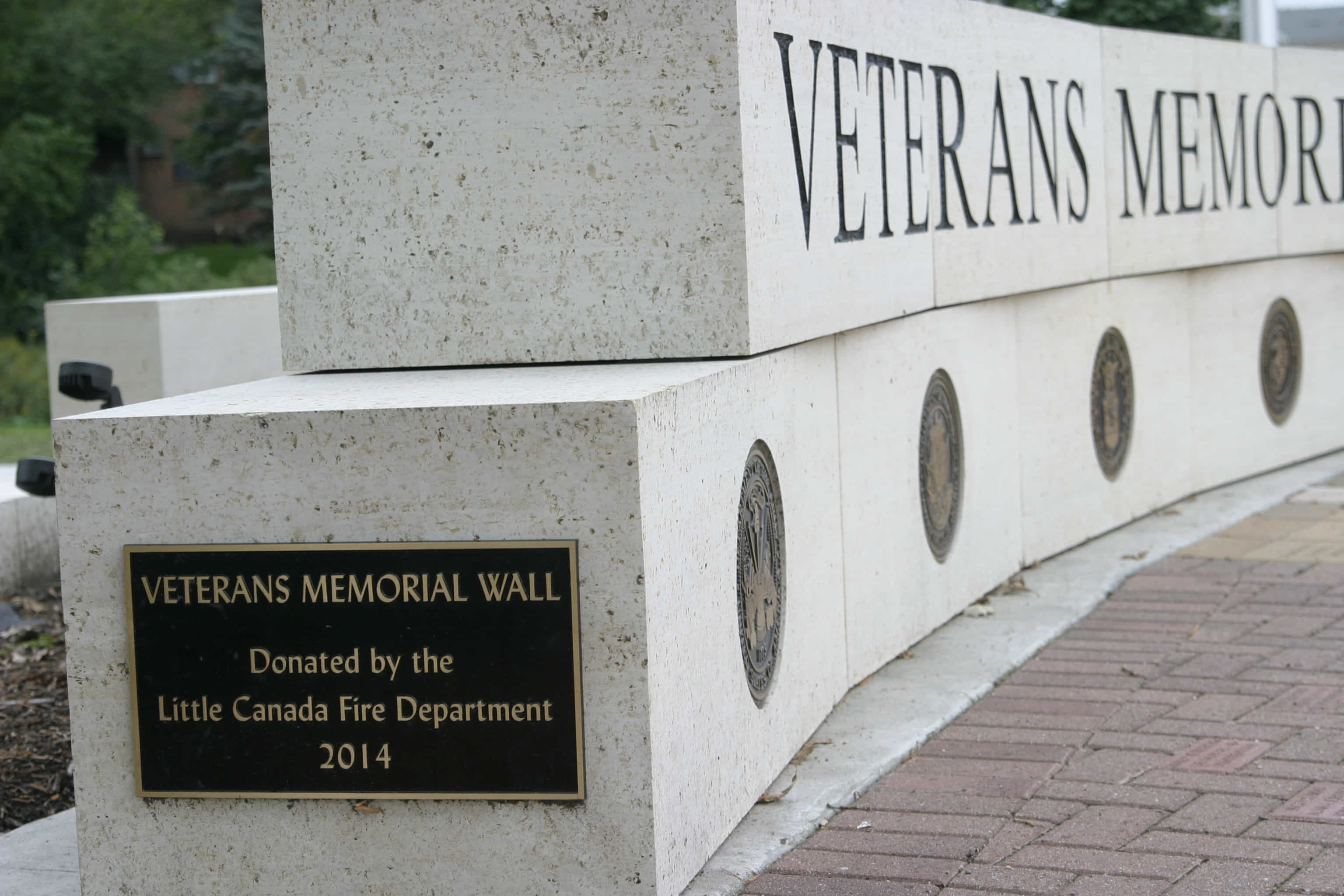 A veterans memorial wall made of two layers of large grey stone blocks. The bottom row contains seals for the branches of the military. The top row has "Veterans Memorial" carved into the stone.